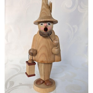 Può includere: Statuina in legno di un uomo con cappello, barba e pipa. La figura tiene in mano una lanterna e una piccola brocca. La statuina è realizzata in legno chiaro con dettagli dipinti sul viso e sugli abiti. La lanterna è rettangolare con una base rosso scuro.