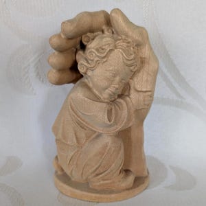May include: A wooden sculpture of a child nestled in a protective hand. The child is wearing a long-sleeved garment and is in a kneeling position. The sculpture is made of light-coloured wood and has detailed carving.