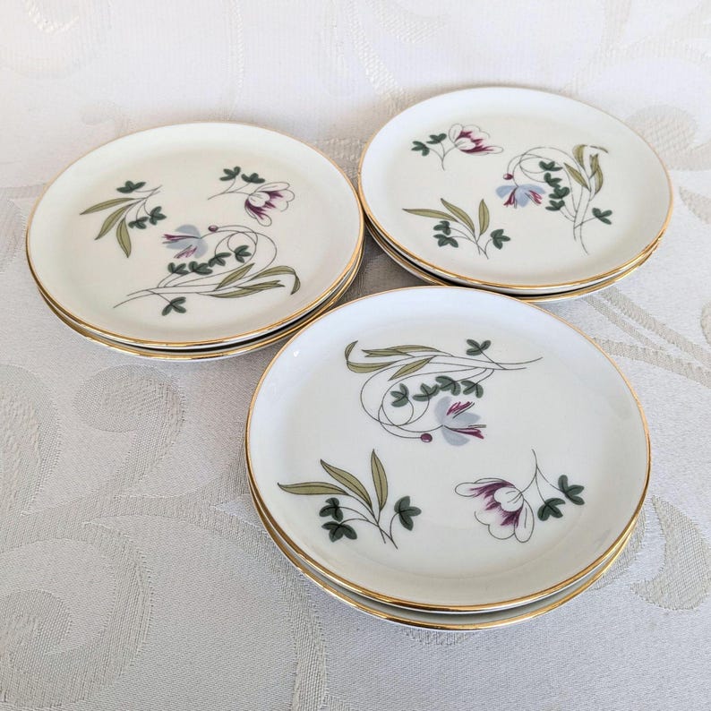 May include: Set of three white ceramic plates with gold rims. Each plate features a floral design with purple, green, and gray accents. The plates are arranged on a white surface.