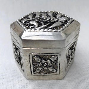 Könnte beinhalten: Eine sechseckige Silberdose mit filigranen Blumendekoren auf allen Seiten. Die Dose ist aus filigranem Metall gefertigt und hat einen Scharnierdeckel.