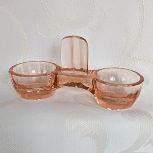 Könnte beinhalten: Ein Vintage-Salz- und Pfefferkeller aus rosa Glas. Das Doppelbecken-Design verfügt über zwei kreisförmige Fächer, die durch eine Brücke verbunden sind, mit einem Griff in der Mitte. Das Glas hat eine zarte, durchscheinende Qualität.