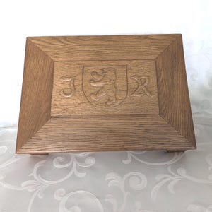May include: A rectangular wooden box with a light brown finish. The top features a carved design with a shield, a lion, and the letters "J R". The box is raised slightly off the surface by small feet.