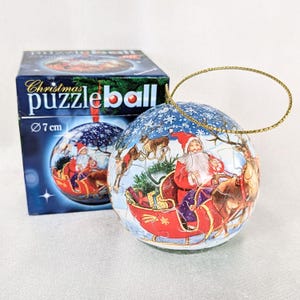 Könnte beinhalten: Eine kugelförmige Weihnachts-Puzzlekugel mit 7 cm Durchmesser, bedruckt mit einer Szene des Weihnachtsmanns im roten Schlitten, der von Rentieren gezogen wird. Die Puzzlekugel hat eine goldene Aufhängeschnur und steht neben der blauen Box.