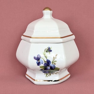 May include: White ceramic lidded jar with a gold trim and a floral design featuring purple violets. The jar has a hexagonal shape and a small gold finial on the lid. A decorative piece for the home.