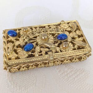 Może przedstawiać: Prostokątne, złote pudełko na biżuterię z misternym filigranem kwiatowym i liściastym. Pudełko posiada kilka owalnych, ciemnoniebieskich kamieni szlachetnych i złote akcenty. Zapięcie to mały, prostokątny element. Pudełko jest zamknięte.