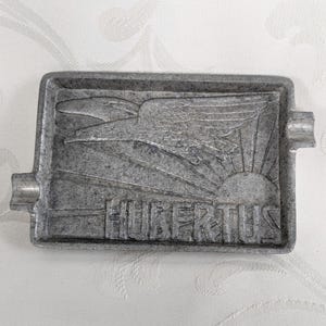 Könnte beinhalten: Ein Aschenbecher aus grauem Metall mit einem erhabenen Design eines Vogels im Flug mit einer aufgehenden Sonne dahinter. Das Wort "Hubertius" ist unter dem Design graviert.
