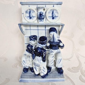 Può includere: Statuina in ceramica blu e bianca raffigurante tre bambini con una barca a vela giocattolo. La statuina ha una mensola con un piccolo vaso e due piatti con disegni di barche a vela. I bambini sono vestiti con abiti da marinaio.