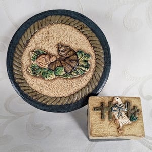 Może przedstawiać: Dwa dekoracyjne elementy ceramiczne. Jednym z nich jest okrągły talerz z reliefem śpiącego kota, otoczony sznurkową obwódką i ciemnoniebieską krawędzią. Drugi to mały prostokątny blok z reliefem gęsi i płotu.