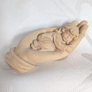 May include: A wooden carving depicting a sleeping baby nestled in a hand. The baby, in a long-sleeved garment, is curled up. The sculpture is crafted from light-toned wood, showcasing intricate details.