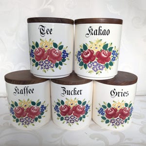 Op de afbeelding: Set van vijf witte keramische potten met houten deksels. Elke pot heeft een bloemenontwerp en een zwart woord aan de voorkant: "Tee", "Kakao", "Kaffee", "Zucker" en "Gries".