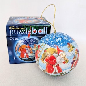 Könnte beinhalten: Eine kugelförmige Weihnachts-Puzzlekugel mit 7 cm Durchmesser, bedruckt mit einer Illustration des Weihnachtsmanns mit Geschenkesack vor einem verschneiten blauen Hintergrund. Die Kugel hat eine goldene Aufhängeschnur und eine blaue Box.