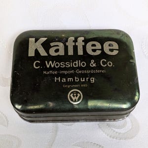 Op de afbeelding: Vintage rechthoekige koffieblik met het woord "Kaffee" in grote, zilveren letters. Het blik bevat ook de tekst "C. Wossidlo & Co., Kaffee-import-Grossrösterei, Hamburg, Gegründet 1885" en een cirkelvormig logo.