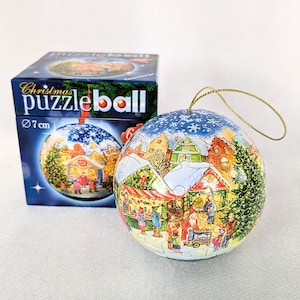 Könnte beinhalten: Eine 7 cm große Weihnachts-Puzzlekugel mit goldener Aufhängeschnur, bedruckt mit einer winterlichen Weihnachtsmarktszene inklusive Menschen und Tannen. Das Produkt steht neben der blauen Kartonverpackung mit der Aufschrift Christmas puzzleball.