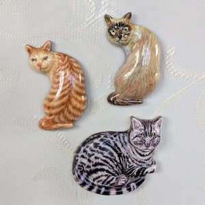 Könnte beinhalten: Drei Keramik-Katzenfiguren in verschiedenen Posen. Eine ist eine orangefarbene Tabbykatze, eine weitere eine Siamkatze und die dritte eine grau-schwarz gestreifte Katze. Die Figuren sind detailliert und scheinen handbemalt zu sein.