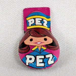 May include: A pink Pez candy dispenser topper featuring a cartoon character with brown hair, wearing a blue and yellow hat with the word PEZ printed in white, and a blue collar with the word PEZ printed in large white letters at the bottom.