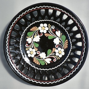 Op de afbeelding: Zwarte decoratieve schaal met een opengewerkte rand en een bloemmotief. De schaal heeft een centrale bloemenkrans in wit, groen, geel en bruin tegen een zwarte achtergrond. De rand heeft een witte, rode en witte rand.