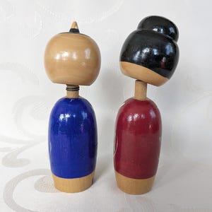 May include: Two wooden Japanese kokeshi dolls, one with a blue and white patterned kimono and the other with a red and white patterned kimono. The dolls have black hair and are painted with traditional Japanese designs.