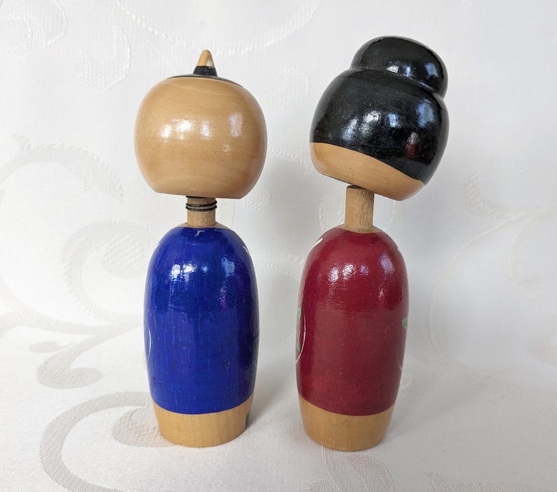 May include: Two wooden Japanese kokeshi dolls, one with a blue and white patterned kimono and the other with a red and white patterned kimono. The dolls have black hair and are painted with traditional Japanese designs.