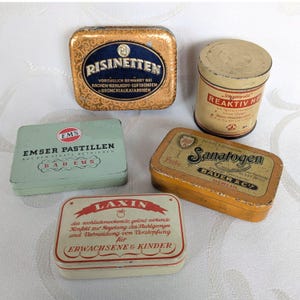 May include: A selection of antique metal tins. The image shows a gold and blue tin with "RISINETTEN", a cylindrical tin with "REAKTIV NE", a green tin with "EMSER PASTILLEN", a yellow tin with "SANATOGEN", and a white tin with "LAXIN".