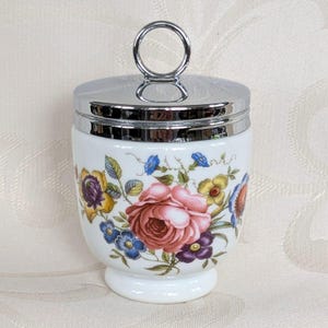 Könnte beinhalten: Weißer Porzellan-Eierbecher mit floralem Design mit rosa Rosen, blauen, gelben und lila Blumen. Der Eierbecher hat einen silberfarbenen Metalldeckel und Griff. Der Sockel ist ein kleiner, abgerundeter Sockel.