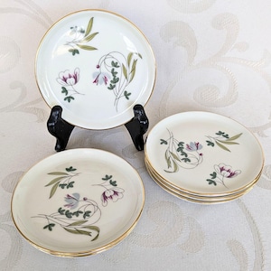 May include: Set of vintage white porcelain plates with gold rims. Each plate features a floral design with purple, green, and gray accents. One plate is displayed on a black stand, while others are stacked. A classic tableware set.