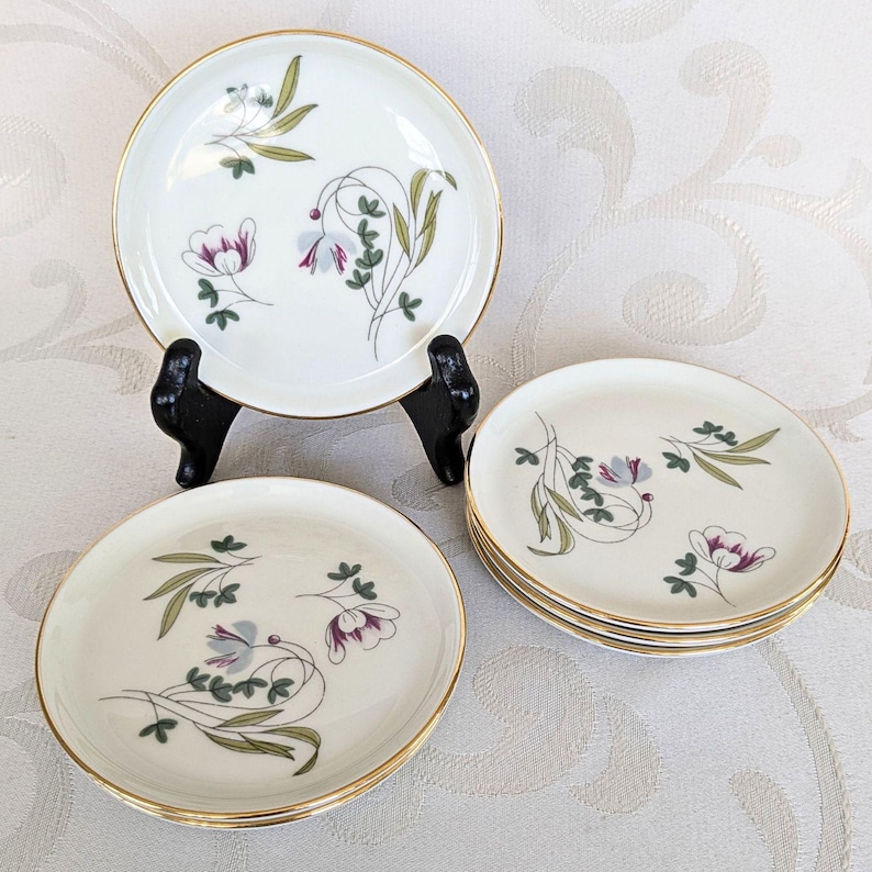 May include: Set of vintage white porcelain plates with gold rims. Each plate features a floral design with purple, green, and gray accents. One plate is displayed on a black stand, while others are stacked. A classic tableware set.