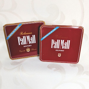 Op de afbeelding: Twee rode en gouden Pall Mall sigarettenkokers. De kokers hebben de woorden "Pall Mall Export" in het wit, met "American Blend" op een blauwe banner. De kokers liggen op een wit oppervlak met een patroon.