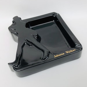 May include: Black ceramic ashtray featuring a raised silhouette of a man in a walking pose. The ashtray is square with rounded corners and a rectangular inset for holding cigarettes. The words "Johnnie Walker" are printed in gold lettering on the bottom right corner of the ashtray.