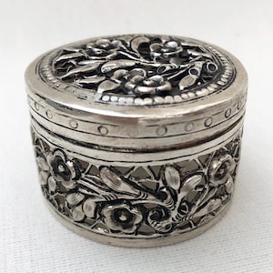 May include: A silver trinket box with intricate floral designs cut out of the metal. The box is round and has a lid.