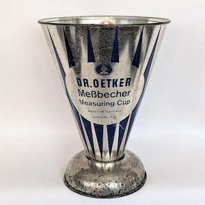 May include: A conical metal measuring cup with a silver finish and blue vertical stripes. The text reads DR. OETKER Meßbecher Measuring Cup, Made in W.-Germany, Artikel Nr. 825. It features a wide circular base and a flared top rim.