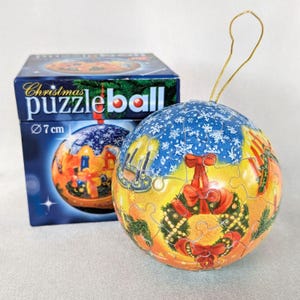 Könnte beinhalten: Eine kugelförmige Weihnachts-Puzzlekugel mit 7 cm Durchmesser, die eine farbenfrohe Weihnachtsszene mit Kranz, Kerzen und Schneeflocken zeigt. Die Puzzlekugel hat eine goldene Aufhängeschnur und steht neben ihrer blauen Kartonverpackung.