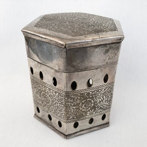 Pode incluir: Uma caixa hexagonal de prata com um design floral intrincado e pequenos furos ao redor dos lados. A caixa tem uma tampa com um design semelhante.
