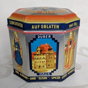 May include: A vintage metal tin with a red, yellow and blue design. The tin is labelled "Lebkuchen auf Oblaten" and "Dürer Haus". It features a picture of a building and two figures. The text on the tin reads "Fine Almond - and "Elisen" - Spiced Cakes".