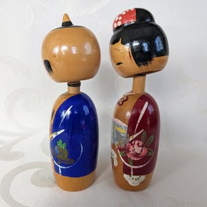 May include: Two traditional Japanese wooden dolls, known as kokeshi, with painted faces and colorful clothing. The doll on the left is wearing a blue kimono with white and green designs, while the doll on the right is wearing a red kimono with white and pink floral designs.