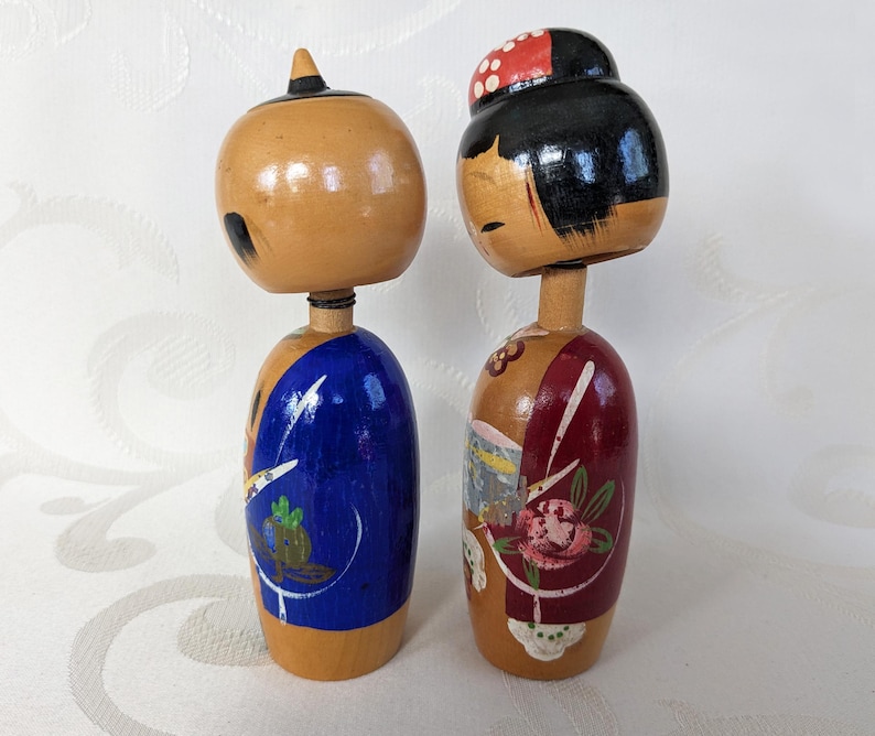 May include: Two traditional Japanese wooden dolls, known as kokeshi, with painted faces and colorful clothing. The doll on the left is wearing a blue kimono with white and green designs, while the doll on the right is wearing a red kimono with white and pink floral designs.