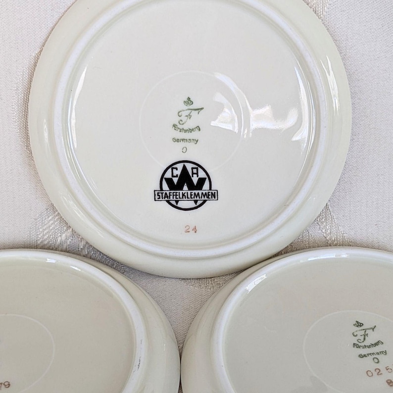 May include: A set of three off-white ceramic plates. Each plate has a raised rim and a central design featuring the text "STAFFELKLEMMEN" and "Germany". The number "24" is printed in red on one plate.
