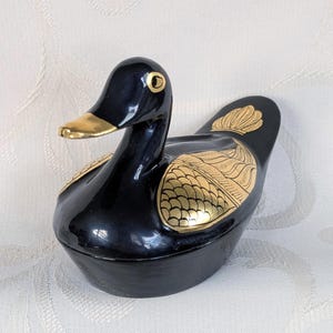Könnte beinhalten: Schwarze Keramik-Entenfigur mit goldenen Akzenten. Die Ente hat eine glänzende Oberfläche, einen goldenen Schnabel und goldene Verzierungen an Flügeln und Schwanz. Ein dekoratives Objekt.