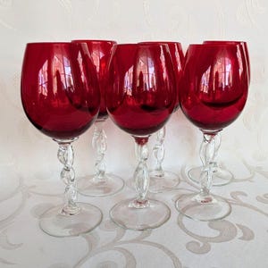 Può includere: Set di sei calici da vino rosso rubino. Ogni bicchiere presenta una coppa arrotondata e un gambo trasparente e ritorto. I bicchieri sono disposti su una superficie bianca a fantasia, riflettendo la luce e aggiungendo eleganza.