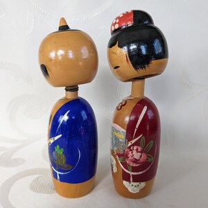 May include: A pair of traditional Japanese wooden dolls, known as kokeshi, with painted faces and colorful clothing. The female doll has a red and white patterned hat and a red and white floral design on her dress. The male doll has a blue and white patterned design on his clothing.