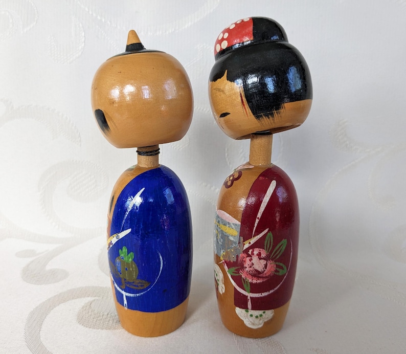 May include: A pair of traditional Japanese wooden dolls, known as kokeshi, with painted faces and colorful clothing. The female doll has a red and white patterned hat and a red and white floral design on her dress. The male doll has a blue and white patterned design on his clothing.