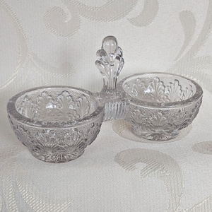 May include: Clear glass double salt cellar with a decorative handle. The two bowls have a detailed floral pattern. The handle is also decorated with a floral design. This vintage piece is perfect for serving salt and pepper.