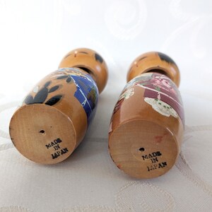 May include: Two wooden Japanese dolls with painted faces and colorful clothing. The dolls are standing on a white surface and the bottoms of the dolls have the text "Made in Japan" printed on them.