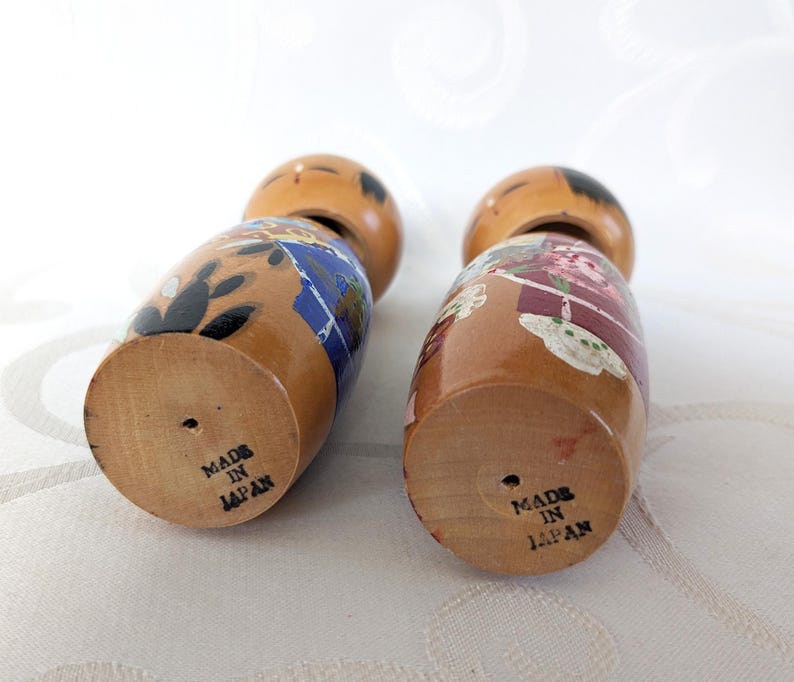 May include: Two wooden Japanese dolls with painted faces and colorful clothing. The dolls are standing on a white surface and the bottoms of the dolls have the text "Made in Japan" printed on them.