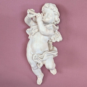 Op de afbeelding: Een crèmekleurige wandplaat met een gebeeldhouwde cherubijn die een fluit bespeelt. De figuur heeft krullend haar, kleine vleugels en is gedrapeerd in vloeiende stof. Het beeldhouwwerk heeft een matte afwerking en een dynamische houding.
