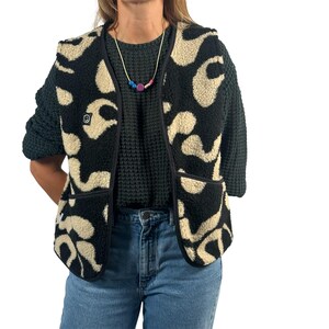 Könnte beinhalten: Eine schwarze und cremefarbene Fleeceweste mit einem einzigartigen Muster. Die Weste hat eine schwarze Paspelierung, zwei Taschen und ein kleines Logo auf der Brust. Sie wird über einem dunkelgrünen Pullover und einer Jeans getragen.