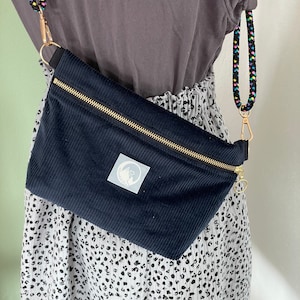 Könnte beinhalten: Eine schwarze Cord-Bauchtasche mit einem goldenen Reißverschluss und einem kleinen weißen Logo. Die Bauchtasche wird über einem weißen Rock mit schwarzen Punkten getragen.