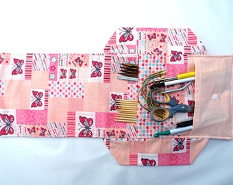 Könnte beinhalten: Ein rosa Stoff-Stricknadel-Etui mit Schmetterlingsmuster. Das Etui ist geöffnet und zeigt verschiedene Strickwerkzeuge, darunter Nadeln, Scheren und Marker. Das Etui ist aus einem Patchwork-Design mit rosa, roten und weißen Farben gefertigt.