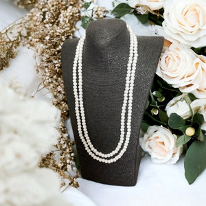 Pode incluir: Um colar de pérolas de duas voltas em um expositor de veludo preto, cercado por flores brancas.