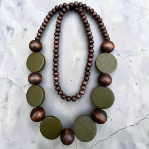 May include: A double-strand necklace featuring a mix of dark brown spherical wooden beads and larger, flat, olive green circular wooden discs. The design alternates between the smooth, stained brown spheres and the matte olive green circular shapes.