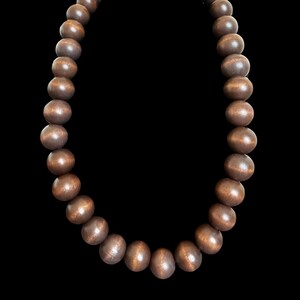 May include: A necklace featuring large, round, wooden beads in varying shades of brown. The beads are strung together, creating a simple yet elegant design. The necklace is displayed against a black background.